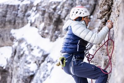 Climber with helmet and rope on a rock face in snow-covered mountains.