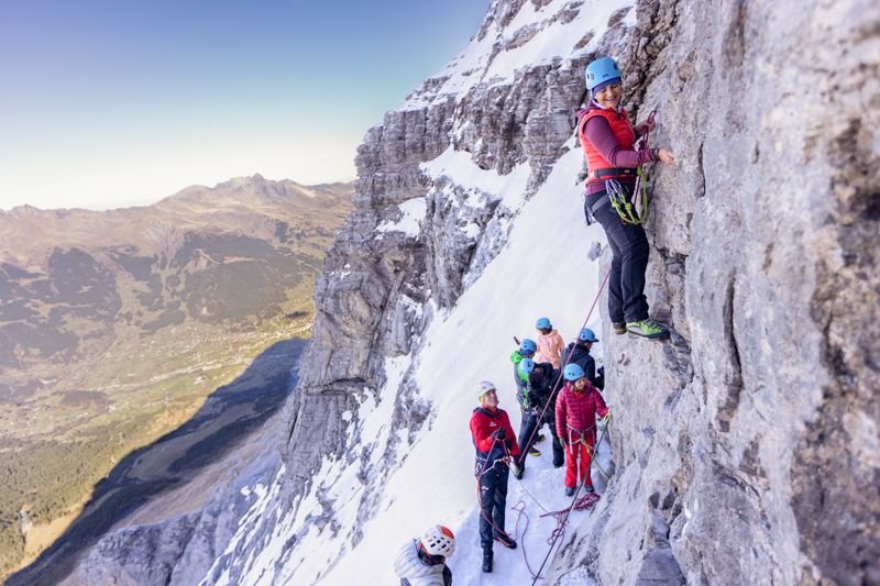 People with helmets and climbing gear on a snow-covered rock face in the mountains.