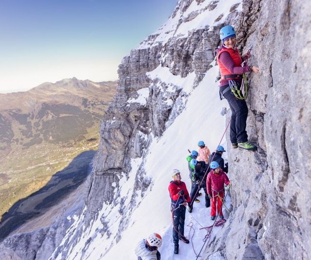 People with helmets and climbing gear on a snow-covered rock face in the mountains.