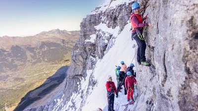 People with helmets and climbing gear on a snow-covered rock face in the mountains.