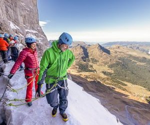 People with helmets and climbing harnesses on a snowy mountain path, secured with ropes.