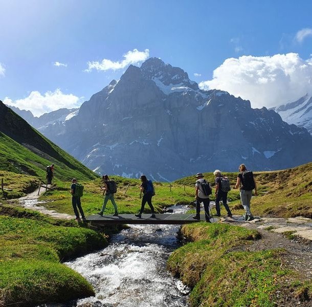 Hikers crossing a stream with a bridge with the Wetterhorn in the background.