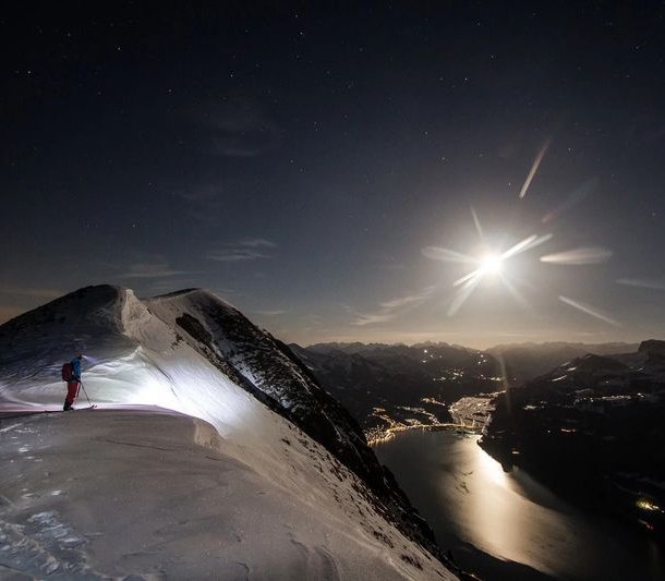 Skier with poles and backpack on snowy mountain ridge at night, overlooking a lake and distant lights.