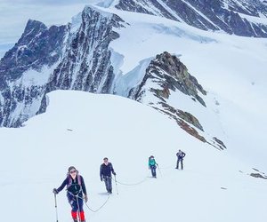 Four climbers with poles and harnesses traverse a snowy mountain ridge, roped together.