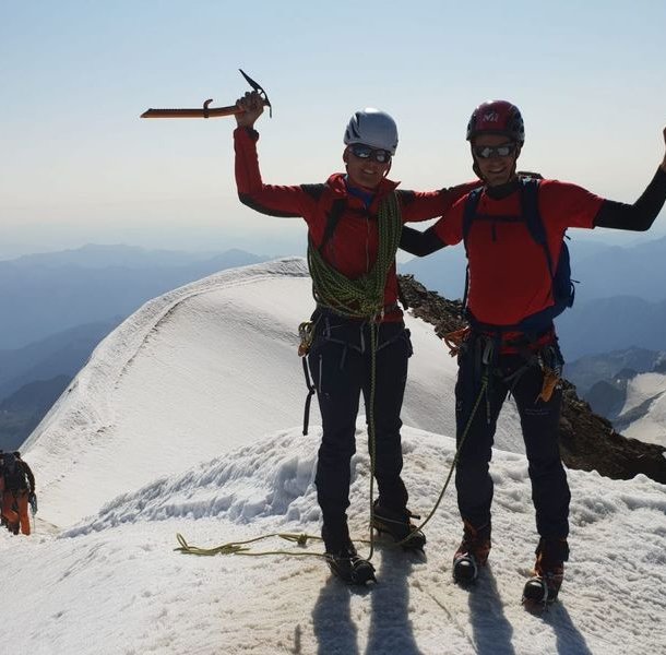 Climbers in helmets and harnesses stand on a snowy mountain ridge, holding ice axes, with others behind.