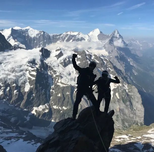 Two climbers with helmets and harnesses on a snowy mountain peak, with a vast alpine backdrop.