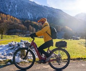 Person cycling on a paved path, wearing a helmet and backpack, with snowy mountains in the background.