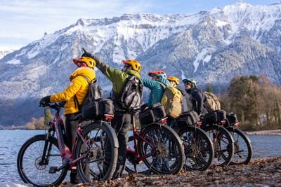 Five people on bikes with helmets and backpacks by a lake, snowy mountains in the background.