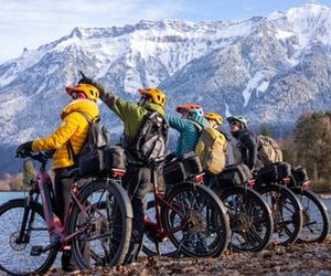 Five people on bikes with helmets and backpacks by a lake, snowy mountains in the background.