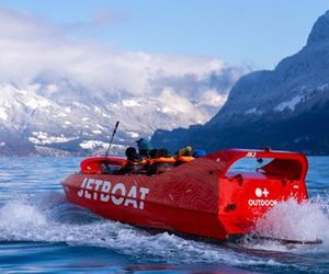 Red jet ski with people in life jackets on a lake, surrounded by snow-covered mountains.
