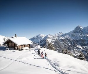 People with ski equipment are hiking in the snow next to a hut in the Swiss Alps.