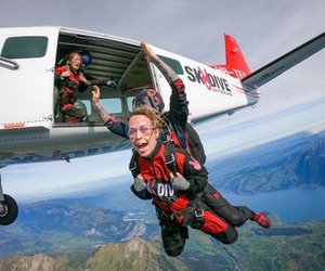 Tandem skydivers in red jumpsuits and goggles exit a plane over mountains and a lake in Switzerland.