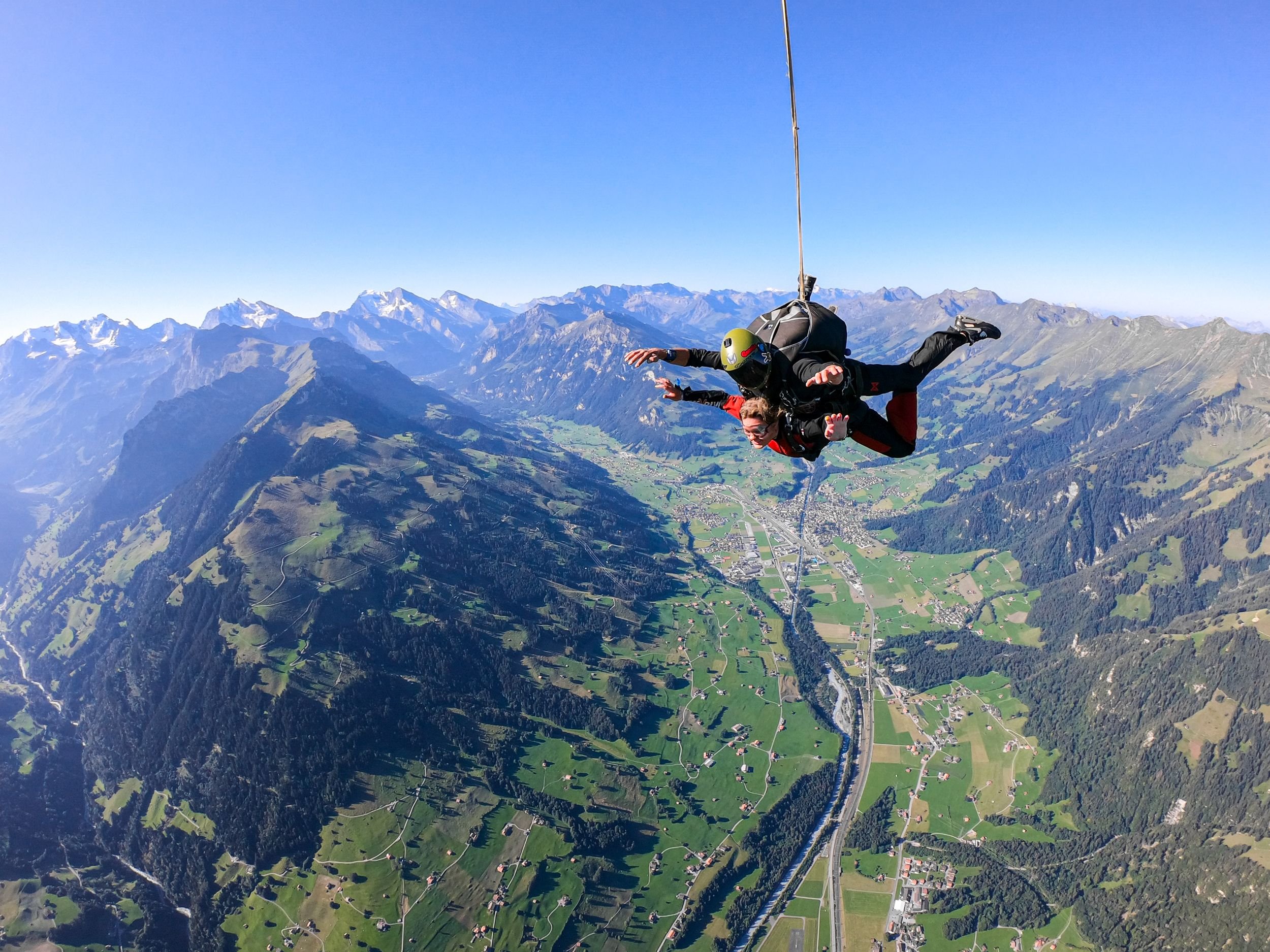 Zwei Personen machen Tandem-Fallschirmspringen über einer bergigen Landschaft, tragen Helme und Gurte.