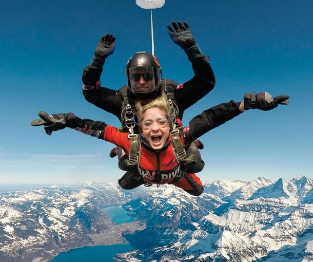 Zwei Personen machen Tandem-Fallschirmspringen über schneebedeckten Bergen, tragen Helme und Gurte.