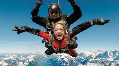 Two people tandem skydiving over snowy mountains, wearing helmets and harnesses.