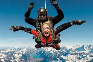 Two people tandem skydiving over snowy mountains, wearing helmets and harnesses.