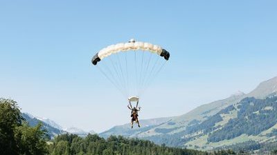 Zwei Personen gleiten mit einem Gleitschirm über einem grasbewachsenen Feld, im Hintergrund Berge.