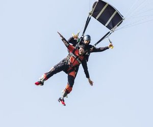 Zwei Personen machen Tandem-Fallschirmspringen mit Fallschirm, tragen Helme und Gurte, vor einem klaren Himmel.