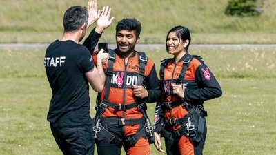 Zwei Personen in Fallschirmspringeranzügen und Gurten stehen auf Gras und interagieren mit einem Mitarbeiter.