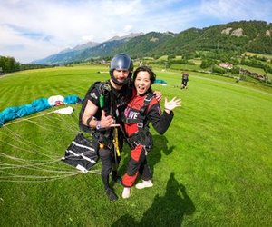 Zwei Personen in Fallschirmausrüstung auf einem grasbewachsenen Feld, mit Bergen im Hintergrund.