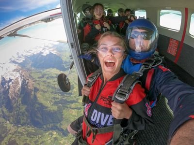 Two people in skydiving gear, including helmets and harnesses, prepare to jump from a plane over mountains.
