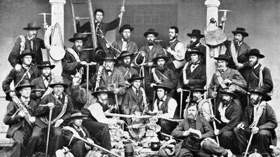 Gruppe von Männern in traditioneller Kleidung mit Wanderstöcken und Ausrüstung vor einem Gebäude.