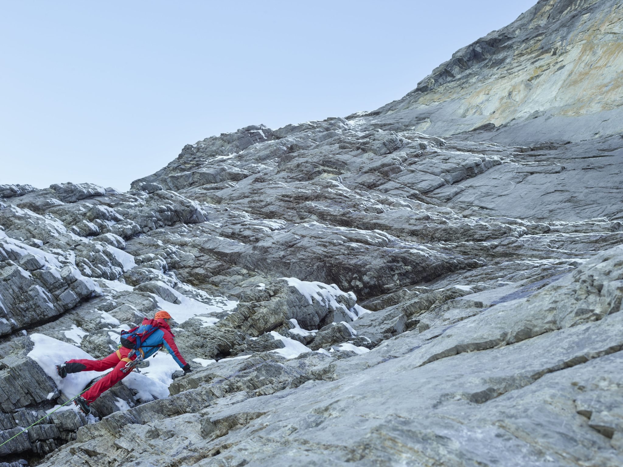 Kletterer mit Helm und Klettergurt auf felsigem Berggelände mit Schnee.