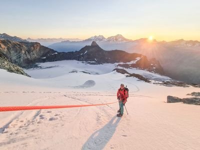 Person with helmet and climbing equipment on a snow-covered mountain at sunrise, mountains in the background.