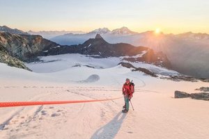 Person with helmet and climbing equipment on a snow-covered mountain at sunrise, mountains in the background.