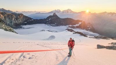 Person with helmet and climbing equipment on a snow-covered mountain at sunrise, mountains in the background.