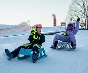 Two people sledding on snow, wearing helmets, goggles, and winter gear, with trees in the background.