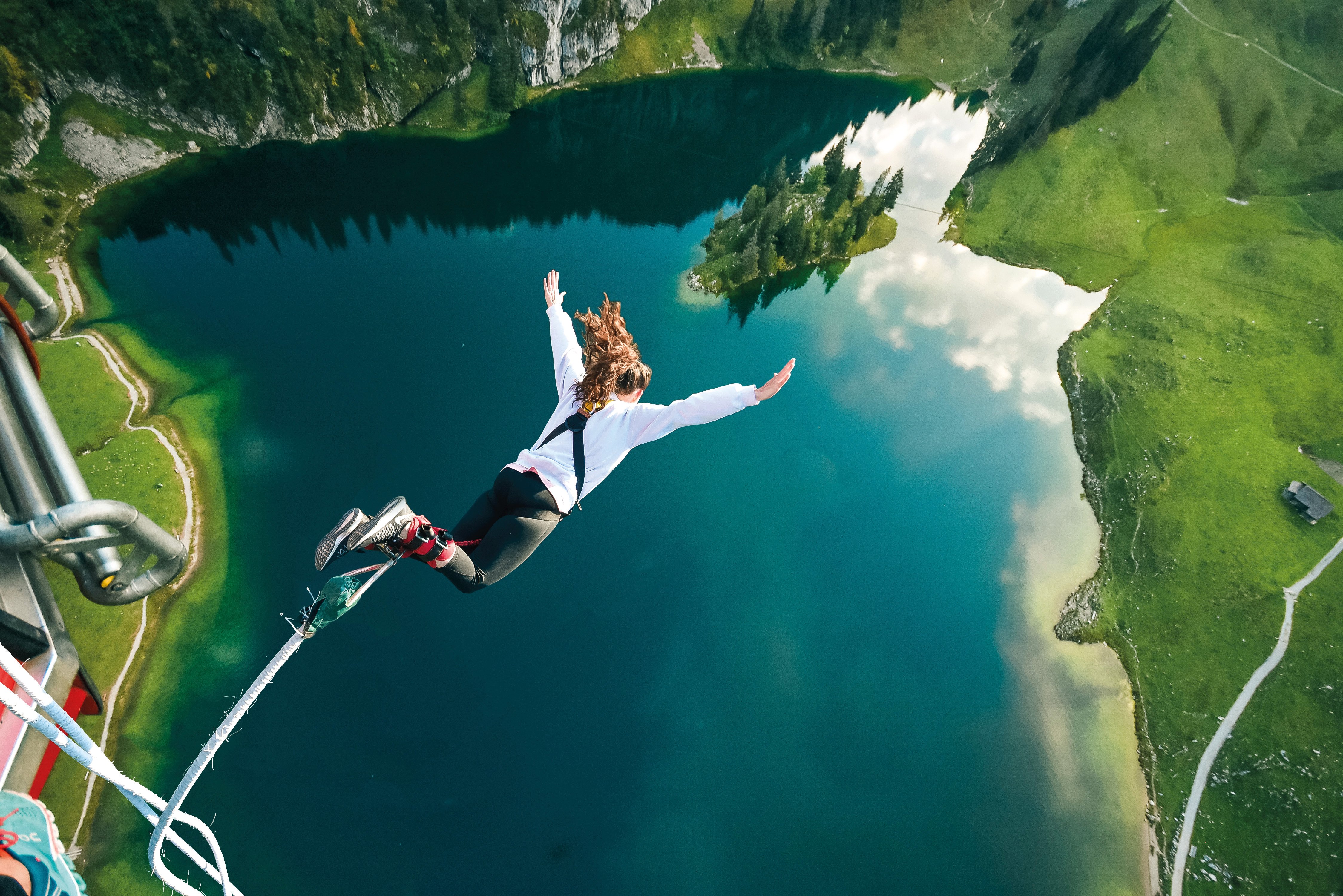 Person beim Bungee-Jumping über einem Bergseeee, trägt ein Sicherheitsgurt und Knöchelriemen, umgeben von Bergen.