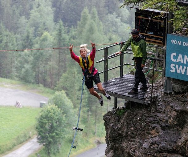 Person im Gurt springt von Plattform für Canyon-Swing; Guide steht in der Nähe; bewaldeter Hintergrund.