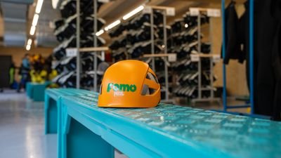 Oranges Kletterhelm mit Aufschrift „Fomo“ auf blauem Metallbank in Ausrüstungsraum mit Regalen voller Schuhe.