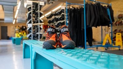 Ein Paar orange-schwarze Wanderschuhe steht auf einer blauen Bank in einem Verleihraum mit Neoprenanzügen und Ausrüstung im Hintergrund.
