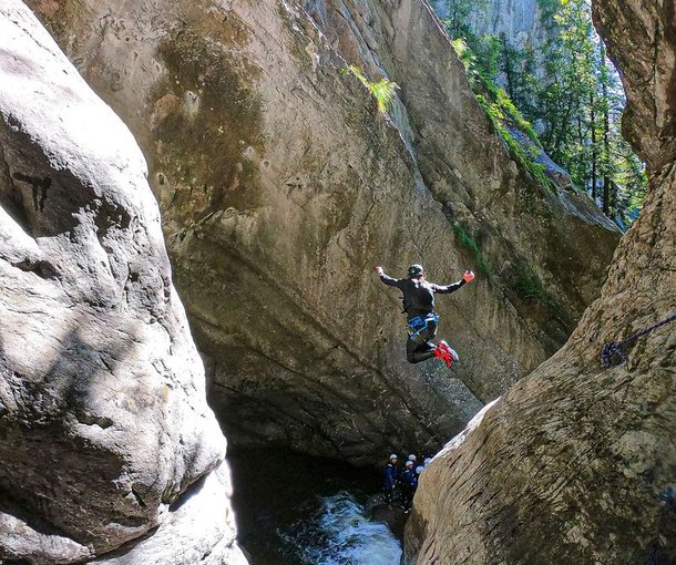 Person in Neoprenanzug und Helm springt in einen Canyon-Pool, während andere unten zuschauen, umgeben von felsigen Klippen.