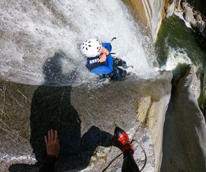 Person in Helm und Neoprenanzug beim Canyoning an einem Wasserfall hinunter, mit sichtbaren Seilen und felsiger Umgebung.