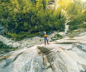 Person rappelling down a steep rock face in a forested canyon, wearing a helmet and harness.