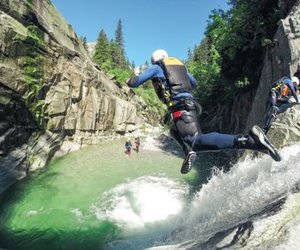 Person in wetsuit and helmet jumps into a canyon pool, others wait on rocks; lush, rocky setting.