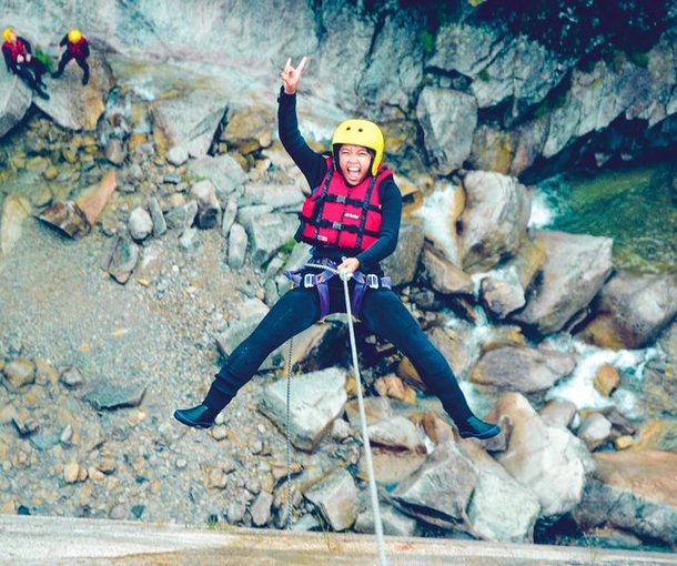 Person in a yellow helmet and red vest rappelling down a rocky canyon, with two others watching.