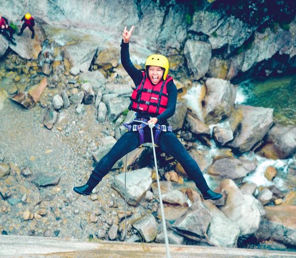 Person in a yellow helmet and red vest rappelling down a rocky canyon, with two others watching.