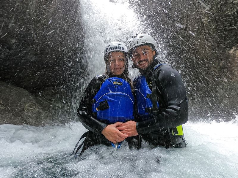 Zwei Personen in Neoprenanzügen und Helmen stehen unter einem Wasserfall in einer felsigen Schlucht und tragen Rettungswesten.