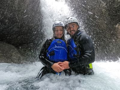Zwei Personen in Neoprenanzügen und Helmen stehen unter einem Wasserfall in einer felsigen Schlucht und tragen Rettungswesten.