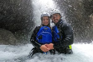 Two people in wetsuits and helmets stand under a waterfall in a rocky canyon, wearing life vests.