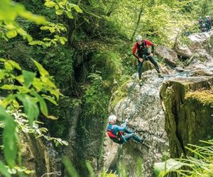 Two people canyoning with helmets and harnesses on a rocky cliff in a lush forest setting.