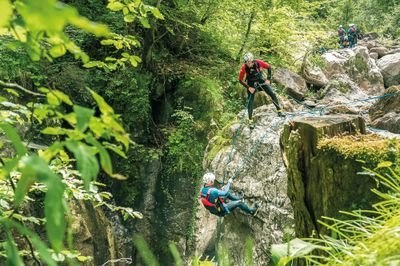 Zwei Personen beim Canyoning mit Helmen und Gurten an einer felsigen Klippe in einer üppigen Waldkulisse.