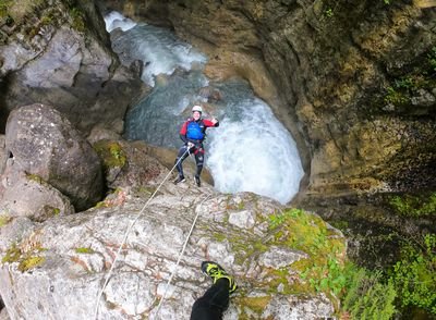 Person in Helm und Neoprenanzug seilt sich an einem felsigen Canyon ab, mit reißendem Wasser darunter.