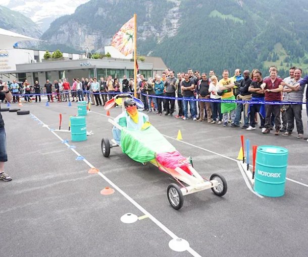 Person in colorful soapbox car on asphalt track, surrounded by spectators in mountain scenery.