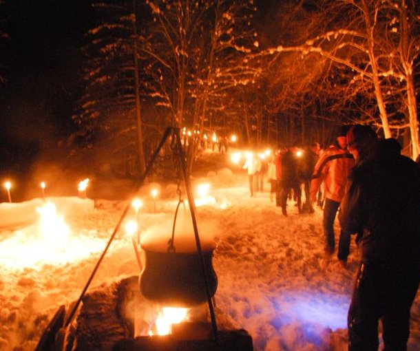 People are walking at night with torches through a snowy forest; campfire and trees in the background.