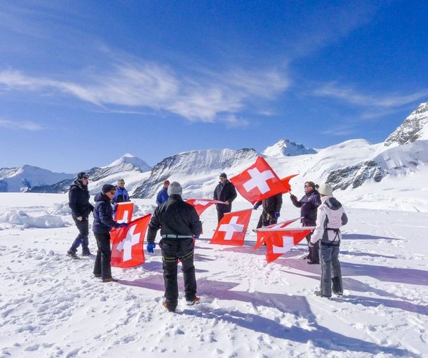 A group with Swiss flags on a snow-covered mountain, some wearing winter clothing and equipment.